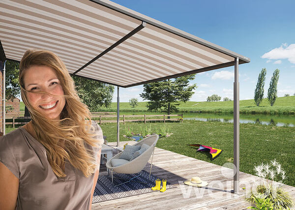 Frau auf Terrasse mit moderner Markise, Gartenmöbeln und Blick auf Wiese und See im Sommer.