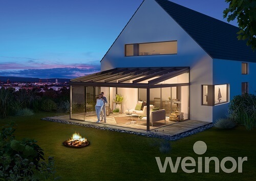 Modernes Einfamilienhaus mit beleuchtetem Wintergarten und Feuerstelle im Garten bei Abenddämmerung.